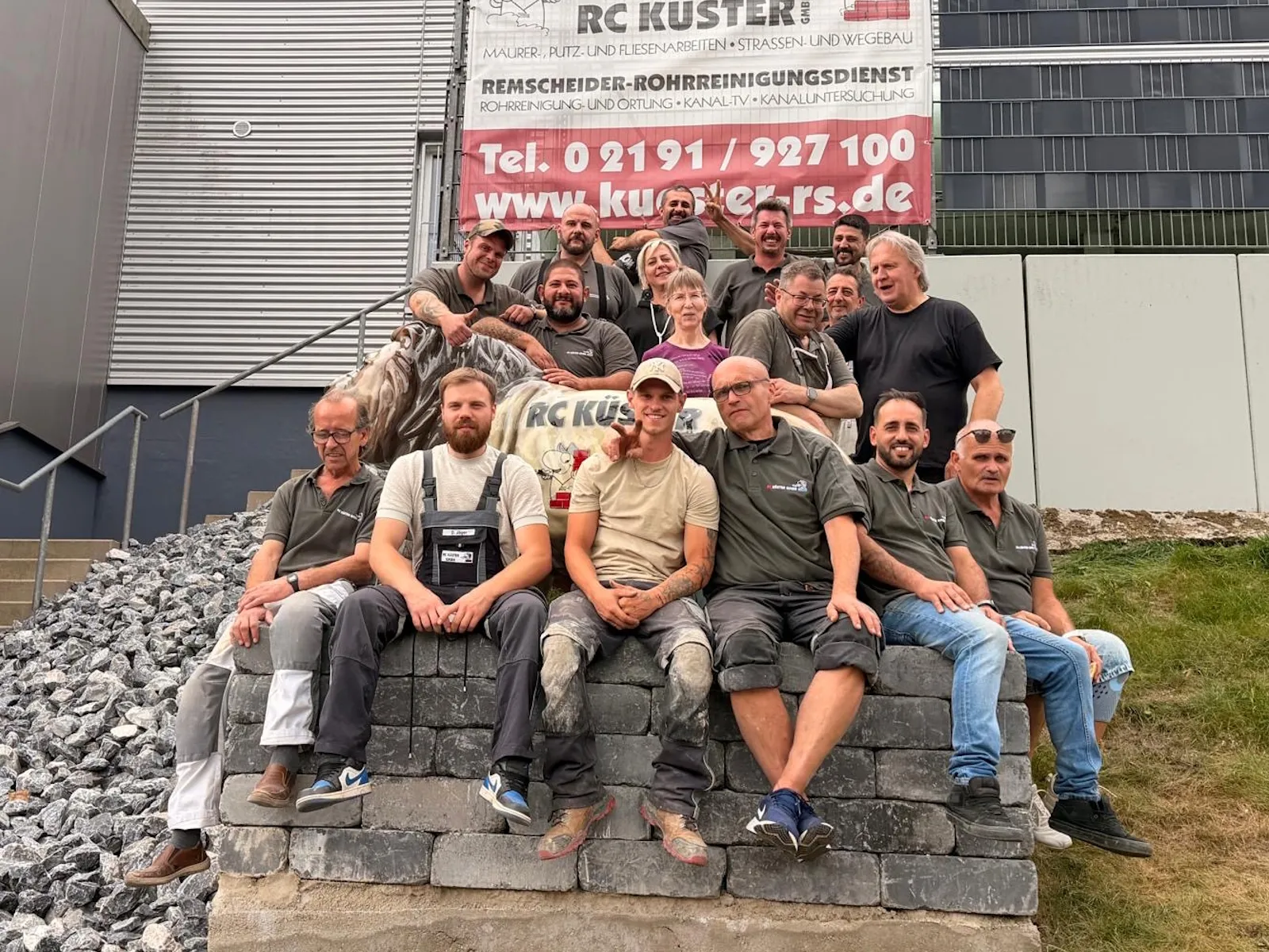Die Belegschaft der RC Küster GmbH auf einer Mauer aus Pflastersteinen vor dem Betriebsgebäude, dahinter ein Banner mit Firmennamen und Telefonnummer